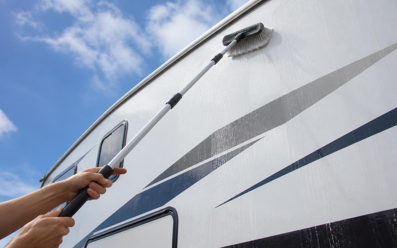 Hands washing the upper side wall of a white motorhome with an extendable brush during RV detailing